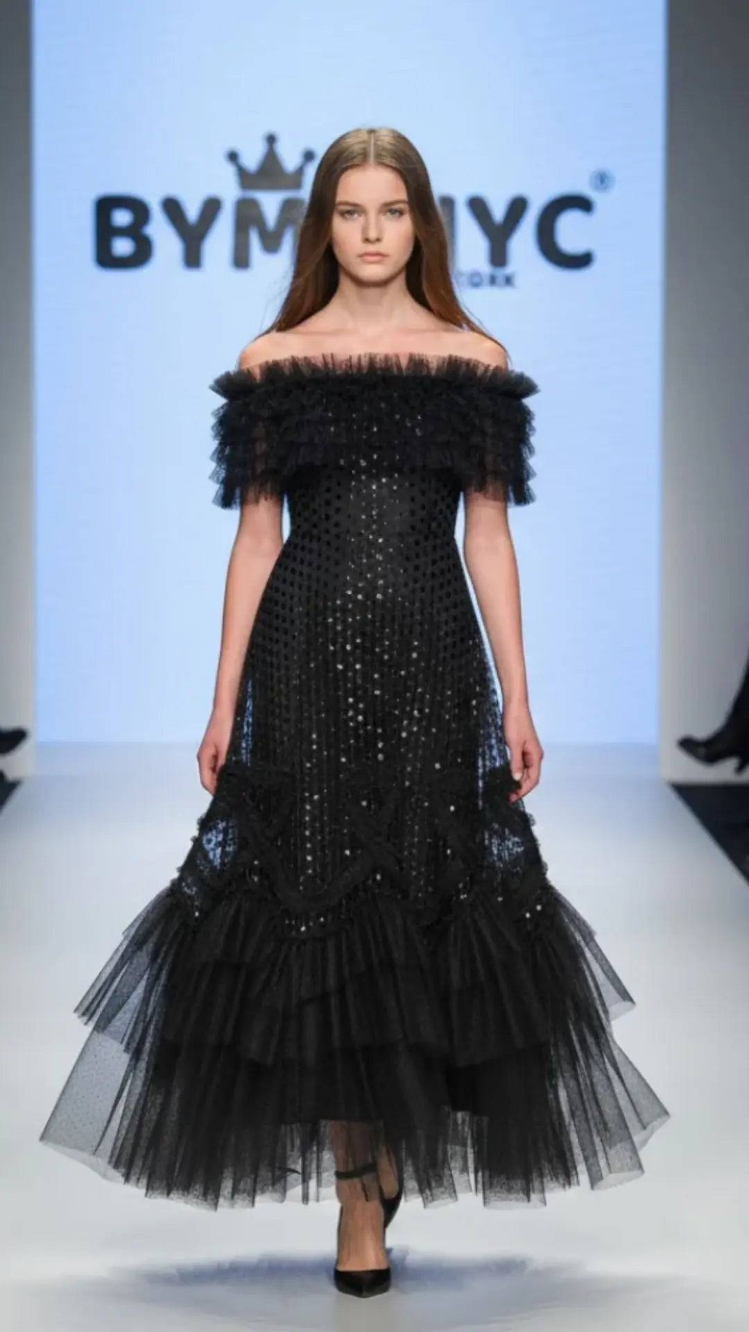 Model wearing a black off-shoulder dress with ruffled layers on a runway with 'BYSNYC' branding.