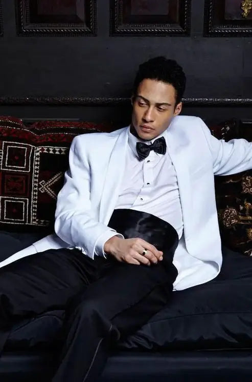Man in a white tuxedo sitting on a dark couch with ornate decorations.